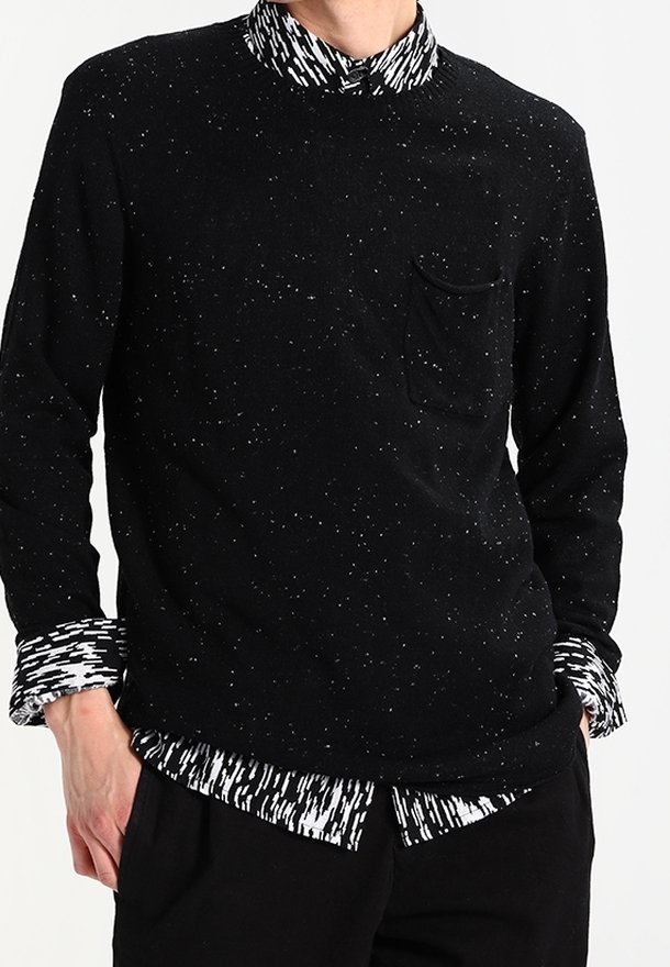 Uomo che indossa un maglione nero a fantasia con un camicia a righe bianca e nera sotto, con colletto e polsini visibili, mani in tasca.