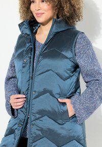 Türkiser, bauschiger Weste mit einem Chevron-Muster, Reißverschluss und Druckknopfverschluss sowie zwei Seitentaschen. Getragen über einem strukturierten blauen Pullover.