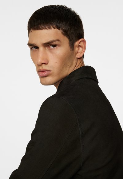 Jonge man met kort bruin haar in een zwarte jas, die over zijn schouder kijkt met een ernstige uitdrukking tegen een effen witte achtergrond.