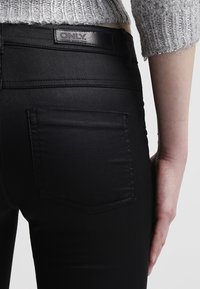 Detailní záběr osoby v černých kalhotách s vysokým pasem s označením "ONLY" a šedém pleteném krátkém svetru, ruka odpočívající po boku.