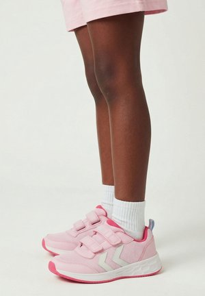 Child wearing pink Velcro sneakers, white socks, and light pink shorts, standing on a plain white surface.