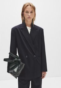 Sacou dublu cu dungi fine bleumarin, cu reverele ascuțite, asortat cu o geantă de mână neagră din piele, cu mânere structurate.