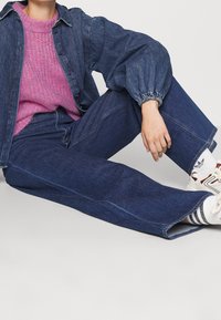 Veste en denim bleu foncé avec fermeture boutonnée sur le devant et poignets élastiques, associée à un pull en maille rose clair, un jean à jambes larges et des baskets à rayures.