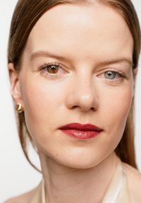 Nahaufnahme einer Person mit zwei unterschiedlich gefärbten Augen, einem braunen und einem blauen. Die Lippen sind in einem tiefen Rot lackiert mit glatter Textur und sie trägt goldene Ohrringe.