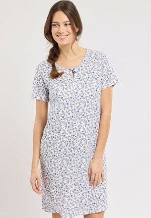 Femme avec une tresse brune portant une robe blanche à manches courtes avec un imprimé floral bleu et un petit nœud à l'encolure, souriante et regardant vers la droite.
