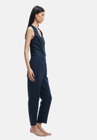 Marineblauwe mouwloze vest met knopen, gecombineerd met loszittende marineblauwe broek. Model draagt minimalistische witte sandalen. Soepele stof, op maat gemaakte pasvorm.