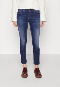 Jeans skinny azul oscuro con un ligero desgaste, diseño de cinco bolsillos, costuras doradas y cierre de botón, combinados con zuecos marrones.