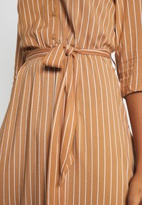 Vestido de rayas marrones con líneas blancas. Presenta un cinturón atado en la cintura, mangas cortas y detalles de botones en el frente. Textura suave.