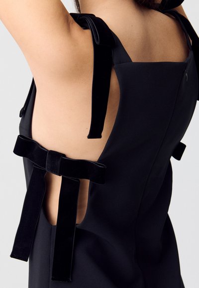 Donna che indossa un vestito senza maniche nero con grandi fiocchi di velluto sulla schiena scoperta e sulle spalline, su uno sfondo semplice.