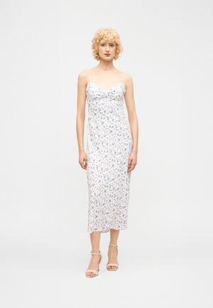 Frau mit lockigem blondem Haar, die ein weißes Midikleid mit blauem Blumenmuster und weiße Schuhe mit Knöchelriemen trägt, steht vor einem schlichten Hintergrund.