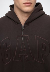 Homme portant un sweat à capuche marron foncé zippé avec des lettres en relief GAP sur la poitrine et la capuche reposant sur les épaules.