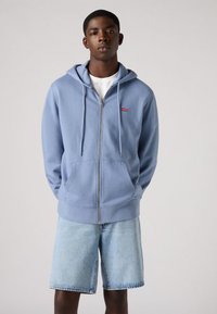 Lichtblauwe ritshoodie van zacht materiaal met een voorzak en trekkoorden. Heeft een klein rood logo op de linkerborst. Gecombineerd met denim shorts.