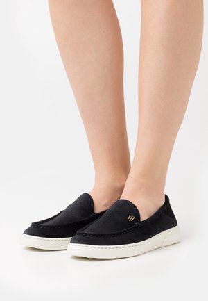 Zapatos slip-on de ante negro con suela de goma blanca, que presentan detalles de costura y un pequeño logo dorado en la parte superior.