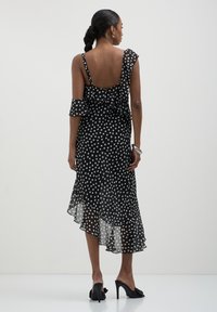 Frau mit geflochtenem Haar, die ein schwarz-weißes, asymmetrisches Kleid mit Punkten und schwarze Heels trägt, steht vor einem einfarbigen Hintergrund.