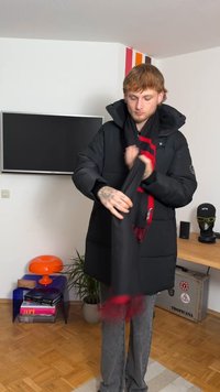 Svart vadderad jacka med huva, med blank yta, stora fickor och en halsduk med röda detaljer. Grå jeans kompletterar outfiten.