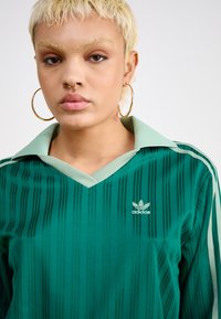 Žalias Adidas polo marškinėliai su vertikaliomis juostelėmis, V formos iškirpte ir šviesiai žaliu apykakle. Mažas Adidas logotipas kairiajame krūtinės pusėje.