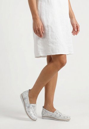 Frau trägt ein weißes knielanges Kleid und silberne, florale Slip-On-Schuhe und steht mit einem Bein über das andere geschlagen.