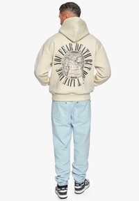 Felpa beige con una grande grafica di un globo e testo sulla schiena, abbinata a jeans azzurri chiari. Sneakers nere completano l'outfit.