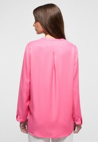 Blusa a maniche lunghe in satin fucsia brillante, con una piega sul retro e orlo arrotondato, con polsini abbottonati e una texture liscia e lucida.