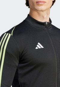 Zwarte sportjack met hoge kraag, ritsluiting en drie groene strepen op de schouders. Voorzien van een wit Adidas-logo op de borst.