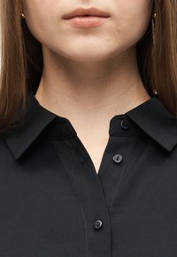 Κοντινό πλάνο του κάτω μέρους του προσώπου και του λαιμού ενός ατόμου που φοράει μαύρο πουκάμισο με γιακά και τα πάνω κουμπιά ξεκούμπωτα, με ορατά ίσια καστανά μαλλιά.