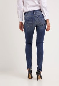 Pantalones vaqueros ajustados de mezclilla azul con una cintura de altura media, que cuentan con dos bolsillos traseros y una textura suave, combinados con zapatos de tacón negro.