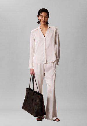 Nő krémszínű hosszú ujjú blúzban és bőszárú nadrágban, nagy sötét színű válltáskát tartva, egy sima szürke háttér előtt áll.