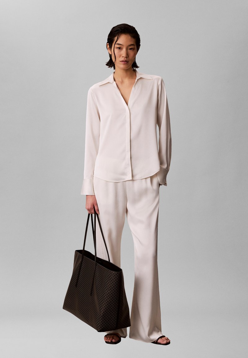 Γυναίκα που φοράει εκρού μακρυμάνικο πουκάμισο και παντελόνι με φαρδιά μπατζάκια, κρατώντας μεγάλη σκούρα τσάντα ώμου, στέκεται μπροστά σε απλό γκρι φόντο.