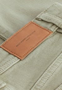 Beež kangas tekstuurilise mustriga, millel on pruun nahklabel, millel on reljeefne kiri "NEXT DENIM COLLECTION EST. 1992." Nähtavad õmblused.