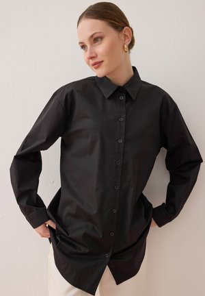 Femme à la peau claire et aux yeux bleus portant une chemise noire boutonnée, debout les mains sur les hanches, regardant sur le côté contre un mur uni.