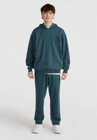 Zeleno-modra kapuca in trenirke iz mehke tkanine, z črnimi napisi. Kapuca ima sprednji žep in kapuco s vrvico.