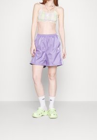 Pantaloncini viola con strisce laterali bianche, un top corto verde chiaro a motivo, e zoccoli verdi. Gli accessori includono una collana multicolore.