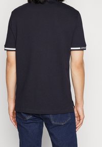 Polo shirt bleu foncé à manches courtes, avec des accents à rayures blanches sur les poignets et une étiquette de logo sur une manche. Tissu lisse au toucher.