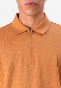 Detailný záber na osobu v štruktúrovanej oranžovej polo košeli na zips s prehnutým golierom na bielom pozadí.