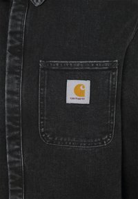 Camisa de mezclilla negra con un bolsillo en el pecho que presenta un parche blanco y un logo amarillo. Diseño clásico con cuello y botones, con una textura sutil.