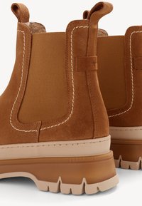 Hnědé semišové Chelsea boty s béžovými masivními gumovými podrážkami a elastickými bočními panely, s poutky na vytažení a bílým kontrastním prošíváním.