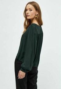 Blusa verde scuro con una texture liscia, maniche lunghe e scollatura arrotondata. Presenta dettagli arricciati sul retro. Abbinata a pantaloni neri.