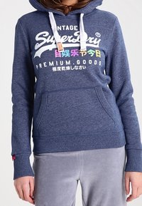Persona che indossa un hoodie blu di Superdry con lacci bianchi e testo multicolore, abbinato a pantaloni grigi chiari.