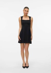 Vrouw die tegen een witte achtergrond staat in een mouwloze zwarte minidress met vierkante halslijn en zwarte puntige hakken.