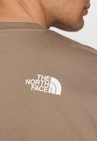 Smeđi majica s teksturom koja sadrži bijeli logo "The North Face" na leđima, s okruglim izrezom i kratkim rukavima.