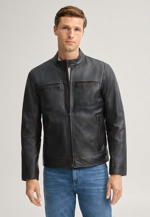 Uomo che indossa una giacca di pelle nera con tasche con cerniera e jeans blu, in piedi davanti a uno sfondo chiaro uniforme.