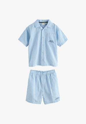 Junger Junge trägt ein blau gestreiftes kurzärmeliges Hemd und passende Shorts, weiße Socken und weiße Turnschuhe, steht auf einem grauen Boden.