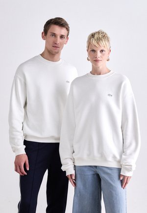 Hvide bomulds sweatshirts med rund hals og ribbede manchetter, der har et lille broderet logo på brystet, parret med afslappede denim og bukser.
