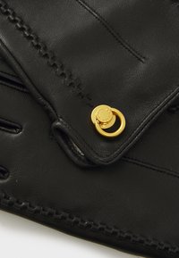 Gant en cuir noir avec des coutures décoratives, un accent en logo doré et un détail en métal circulaire près du poignet.