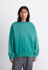 Overdimensioneret teal sweatshirt i blødt stof med ribbede manchetter og kant, der har en rund halsudskæring. Båret over en blå skjorte.