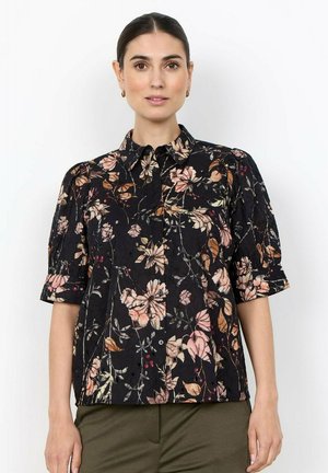 Vrouw draagt een zwart bloemetjesoverhemd met knopen, roze en bruine bloemen en halflange mouwen, gecombineerd met olijfgroene broek.