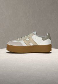 Baskets compensées blanches et beiges avec semelle en gomme, logo "H" beige sur le côté, bout en suède beige et languette beige au talon sur fond neutre.