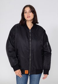 Kobieta w czarnej bomberce z zamkiem z przodu, prążkowanymi mankietami i małą pomarańczową metką przy dolnym wykończeniu, w połączeniu z niebieskimi dżinsami.