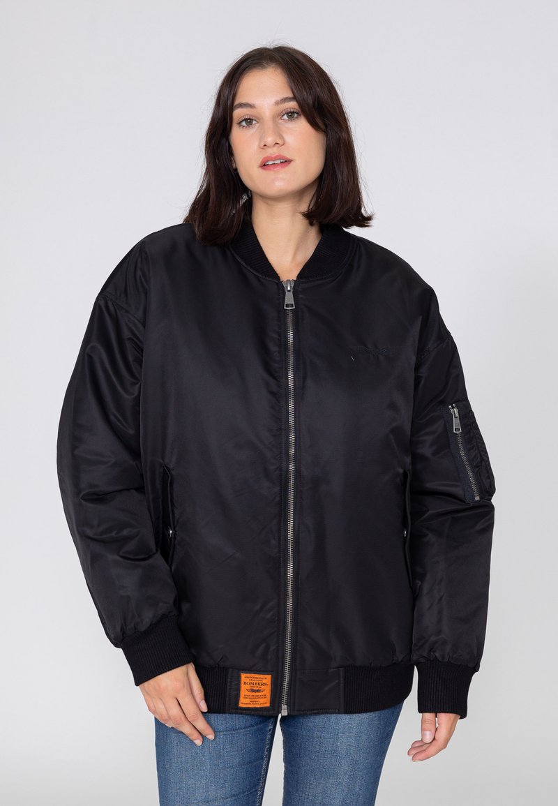 Kobieta w czarnej bomberce z zamkiem z przodu, prążkowanymi mankietami i małą pomarańczową metką przy dolnym wykończeniu, w połączeniu z niebieskimi dżinsami.