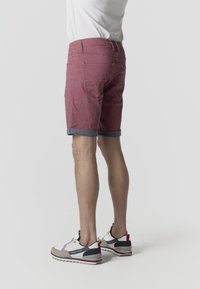 Mann trägt hochgekrempelte burgunderrote Shorts, weißes T-Shirt und beige Sneaker, steht mit sichtbaren Beinen und unterem Oberkörper vor einfachem Hintergrund.